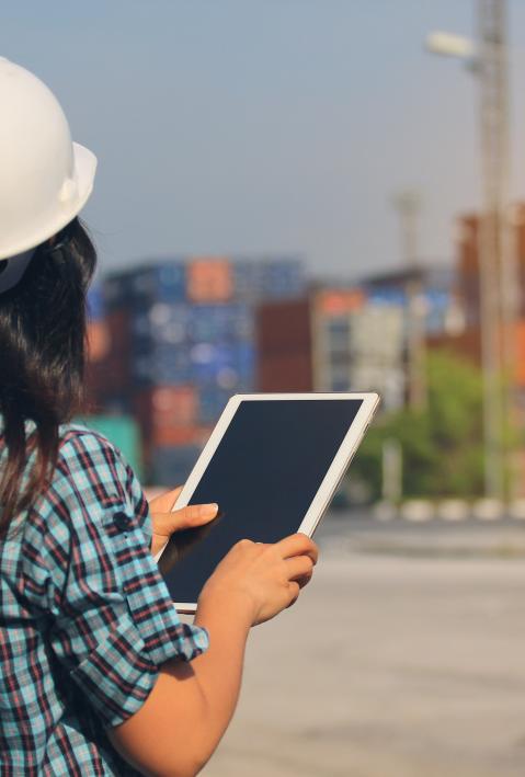 Person mit Helm und Tablet schaut auf Container in der Ferne