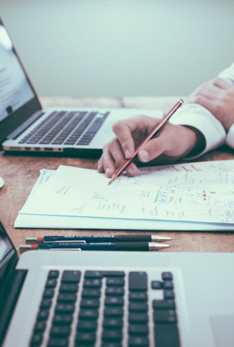 Zwei Personen &uuml;berpr&uuml;fen Daten auf dem Laptop und ausgedruckten Dokumenten am Schreibtisch. 