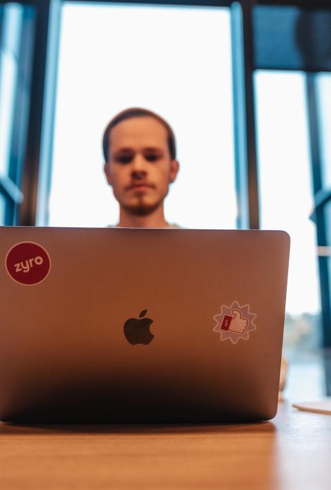 A man sits behind a large wooden table and looks at his Macbook. 